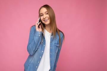 Photo of Attractive sexy positive young blonde woman wearing casual blue jean shirt isolated over pink background holding in hand and talking on mobile phone looking at camera