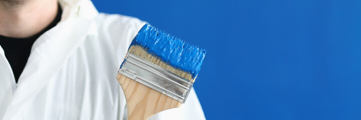 Wall Mural - Man in white protective suit hold paintbrush in his hand. Wooden brush smeared with blue paint against blue wall.