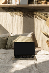 Wall Mural - Blank screen laptop in bed with pillows and linens. Boho style home interior design with sunlight shadows on the wall. Copy space mockup template. Front view freelancer, blogger work business concept.