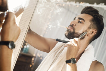 Wall Mural - Man looking in the mirror, smoking a cigarette and shaving his beard with a straight razor