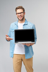 Concentrated young bearded man wearing glasses dressed in jeans shirt holding laptop isolated over grey studio background