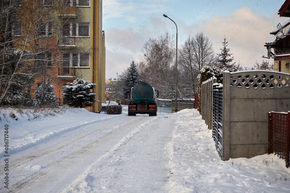 A septic tank, a septic tank that drives along a snowy street, passes a house and an apartment block in the city during winter - obrazy, fototapety, plakaty 