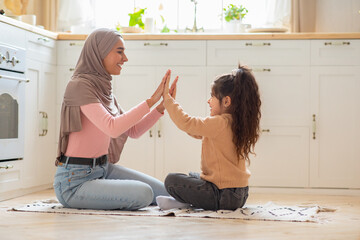 Happy muslim mom playing with her little daughter on floor in kitchen