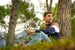 Handsome young man sitting in a forest leaning on a pine tree admiring the landscape with radiant sunlight, man wears tricolor sweater and jeans