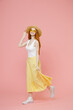 Full length body young smiling redhead ginger woman in straw hat glasses summer clothes maxi skirt walk going waving hand greet someone look camera isolated on pastel pink background studio portrait
