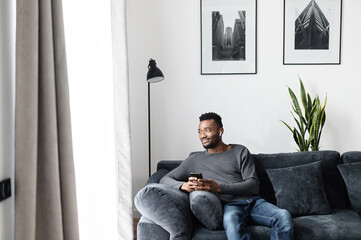 An African-American man spends leisures time with a smartphone at home, thoughtful guy sits on the sofa in cozy living room with a gadget in the hands