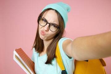 Wall Mural - Close up confused woman student in blue t-shirt hat glasses backpack hold notebooks doing selfie shot on mobile phone isolated on pink background. Education in high school university college concept.