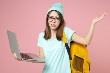 Wall Mural - Confused young woman student in casual blue t-shirt hat glasses backpack work on laptop pc computer spreading hands isolated on pink background. Education in high school university college concept.