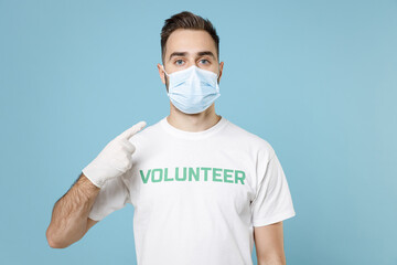 Wall Mural - Young man in volunteer t-shirt sterile gloves pointing finger on face mask to safe from coronavirus virus covid-19 isolated on blue background. Voluntary free work assistance aid help support concept.