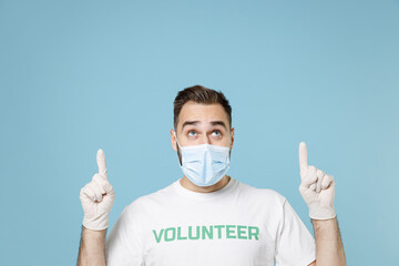 Wall Mural - Young man in volunteer t-shirt gloves face mask to safe from coronavirus virus covid-19 pointing index fingers up isolated on blue background. Voluntary free work assistance aid help support concept.