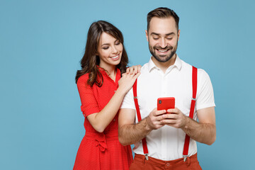 Wall Mural - Smiling young couple two friends man woman wearing white red clothes using mobile cell phone typing sms message isolated on blue color background studio portrait. St. Valentine's Day holiday concept.