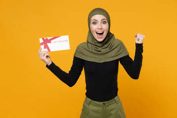 Wall Mural - Excited young arabian muslim woman wearing hijab black green clothes hold gift certificate doing winner gesture isolated on yellow color background studio portrait. People religious lifestyle concept.