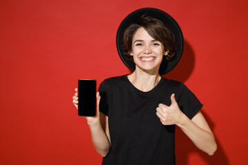 Funny young brunette woman 20s wearing casual basic black t-shirt hat standing hold mobile cell phone with blank empty screen showing thumb up isolated on bright red color background studio portrait.