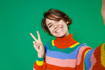 Close up of smiling funny young brunette woman 20s in casual colorful sweater standing doing selfie shot on mobile phone showing victory sign isolated on bright green color background studio portrait.