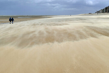 Two people walking on the empty beach with strong sand drifts on a stormy day at low tide; Katwijk, Netherlands, Europe