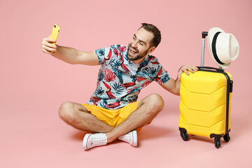 Wall Mural - Full length of cheerful young traveler tourist man sit on floor near suitcase doing selfie shot on mobile phone isolated on pink background. Passenger travel on weekend. Air flight journey concept.