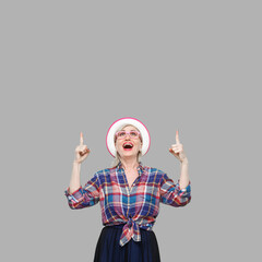 Portrait of surprised modern stylish mature woman in casual style with hat and eyeglasses standing, amazed looking and pointing at up side of wall. indoor studio shot isolated on gray background.