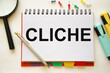 the word cliche is written on a white pad that lies on a white table near a magnifying glass of markers and multi-colored buttons