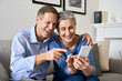 © insta_photos - Happy older mature 50s senior retired couple holding smartphone enjoying using mobile social media apps technology, video calling, shopping in cell phone application sitting on couch at home.