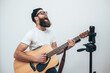 © San4ezz007 - A young stylish guy with a beard wearing casual or music teacher playing guitar in front of smartphone camera. Online guitar training or vocal lesson