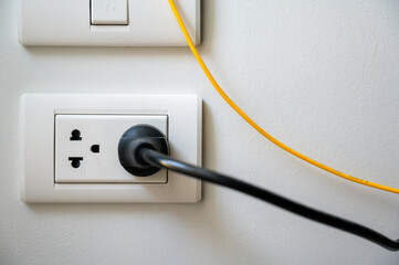 Electric outlet with black electric power cord cable plugged into the wall outlet socket on white wall in the house.