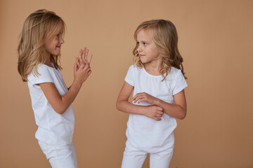 Front portrait of adorable little twin sisters with wavy blond hair, talk each other, wears white clothes, over beige background.