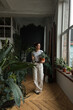 © tantin_s - Young woman gardener holding houseplant in her hands, surrounded by plants and pots. Love plants, hobby, home gardening concept, freelance, small business.