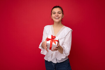 Beautiful happy young brunette woman isolated over colourful background wall wearing stylish casual clothes holding gift box and looking at camera