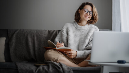 Wall Mural - Beautiful young girl student studying at home online while sitting in warm sweater on the couch