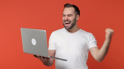 Excited happy bearded young man 20s in white casual t-shirt isolated on orange color background studio. People lifestyle concept. Working on laptop pc computer doing winner gesture screaming say yes