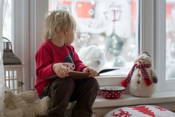 Wall Mural - Cute blond toddler child, sitting on the window, looking at the snow falling outside