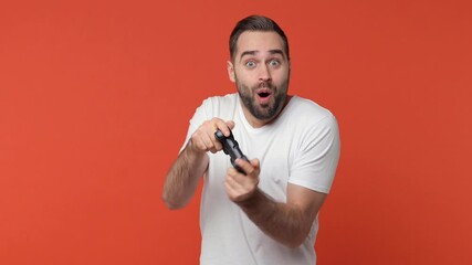 Poster - Excited cheerful bearded young man 20s in white casual t-shirt isolated on orange color background studio. People emotions lifestyle concept. Play pc game with joystick console doing winner gesture