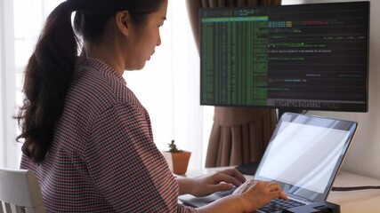 Rearview of young Asian woman, female programmer, data analyst or hacker working with multiple display computer, typing, coding, looking at monitor screen. Indoors, medium shot, work from home concept