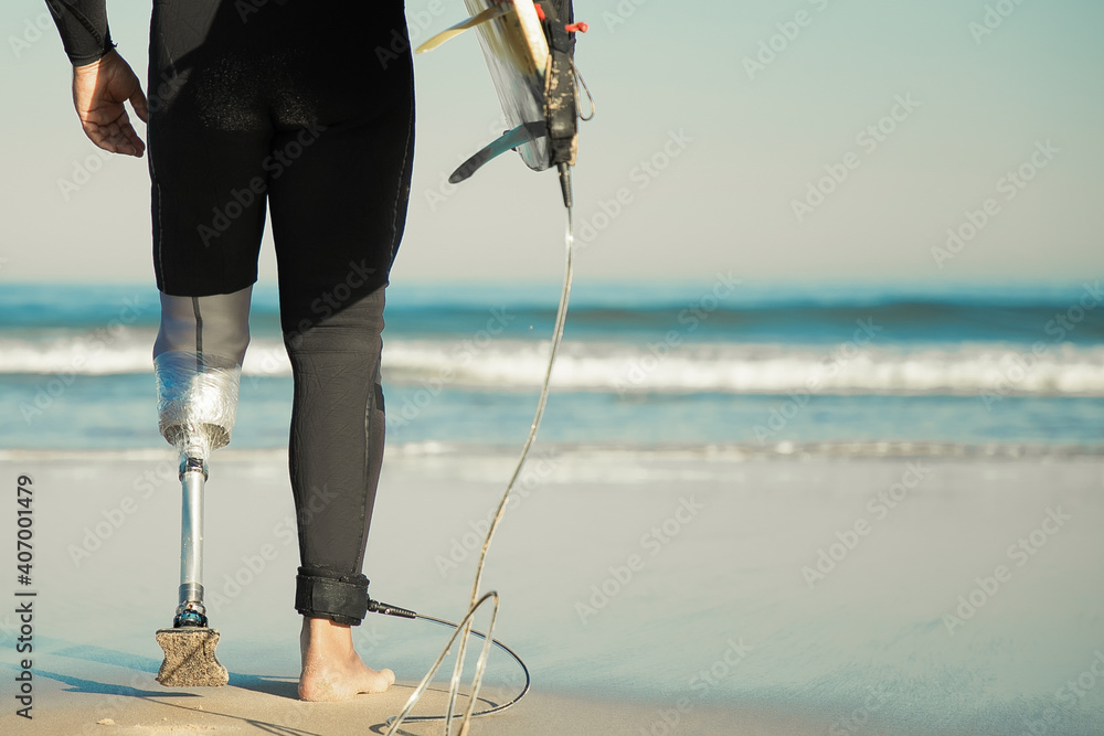 Unrecognizable disabled surfer going to water and carrying longboard. Cropped strong man wearing sportive swimsuit and holding surfboard. Physical disability, surfing and extreme sport concept - obrazy, fototapety, plakaty 