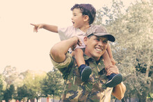 Exited Little Boy Sitting On Dad Neck And Pointing Away. Caucasian Father Holding Son Legs, Smiling, Wearing Army Uniform And Walking In Park. Family Reunion, Fatherhood And Returning Home Concept