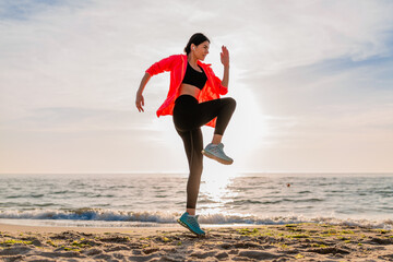 young smiling attractive slim woman doing sports in morning sunrise jumping on sea beach in sports wear, healthy lifestyle, listening to music on earphones, wearing pink windbreaker jacket, having fun