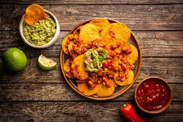 Wall Mural - Delicious plate of yellow corn nachos chips with cheese, minced meat and red hot spicy salsa over wooden table, top view