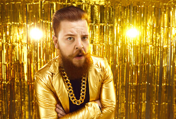 Portrait of eccentric man in extravagant disco party style jacket with gold chain around neck looking at camera with sceptical expression of mistrust on face, standing against shiny golden background