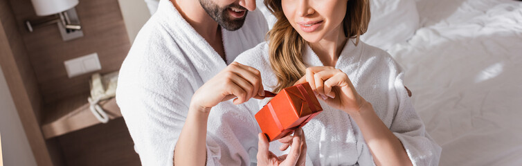 Cropped view of smiling man in bathrobe holding ribbon of present near girlfriend in hotel, banner