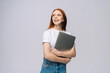 © dikushin - Happy young woman student holding laptop computer and looking away on isolated gray background. Pretty lady model with red hair emotionally showing facial expressions in studio, copy space.