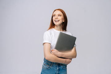 Wall Mural - Happy young woman student holding laptop computer and looking away on isolated gray background. Pretty lady model with red hair emotionally showing facial expressions in studio, copy space.