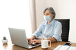© Vadim Pastuh - Senior mature businesswoman with face mask indoors in home office, having video call and typing on the laptop, talking and consulting business client online, writing an email, doing online shopping