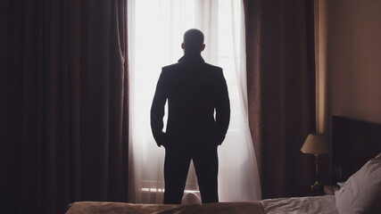 Wall Mural - Portrait handsome young man in an elegant suit standing at window in hotel room. Wedding morning for male. Cute guy is getting ready to meet bride. Concept of happiness and luxury married. Copy space