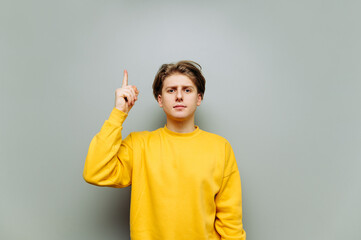 Handsome young man in yellow casual clothes showing thumbs up on copy space and looking at camera with serious face. Isolated on gray background.