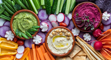 Top Down Close Up View Of Three Colourful Hummus Dips Surrounded By Various Cut Vegetables, Pita Bites And Breadsticks.