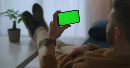 man is holding smartphone horizontal and viewing video, resting at home or office, green screen technology for montage