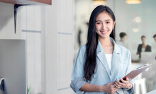 Portrait Of Young Asian Woman In Casual Wear Holdng Clipboard, Smiling And Looking At Camera While Standing In Modern Office Room.