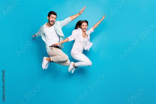Photo of childish carefree young lovers dressed casual shirts jumping high arms hands sides isolated blue color background