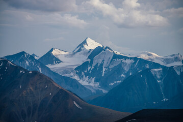 Awesome mountains landscape with big snowy mountain pinnacle in blue white colors under cloudy sky. Atmospheric highland scenery with high mountain wall with pointed top with snow under white clouds.