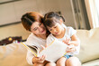 © tuastockphoto - Asian pretty mother and her happy daughter reading story book together on cozy sofa in living room in a homeschooling day they spend a quality time together. Asian family, happy family concept